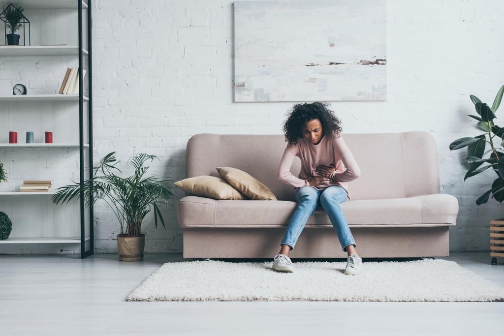 giovane donna afroamericana che soffre di dolore addominale mentre si siede sul divano in soggiorno
 - Foto, immagini