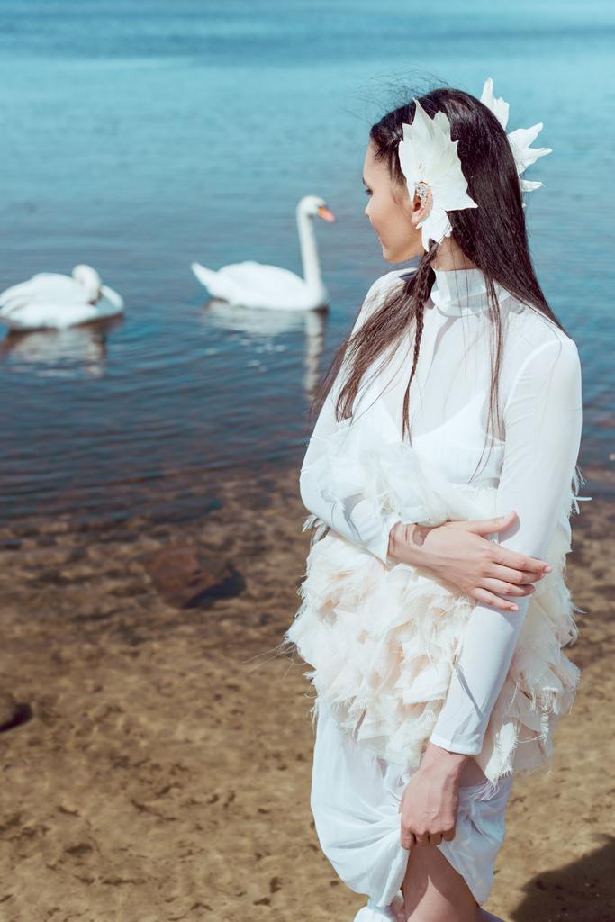 brunetka Kobieta w białym łabędź kostium stojący w pobliżu stawu, patrząc na ptaki - Zdjęcie, obraz