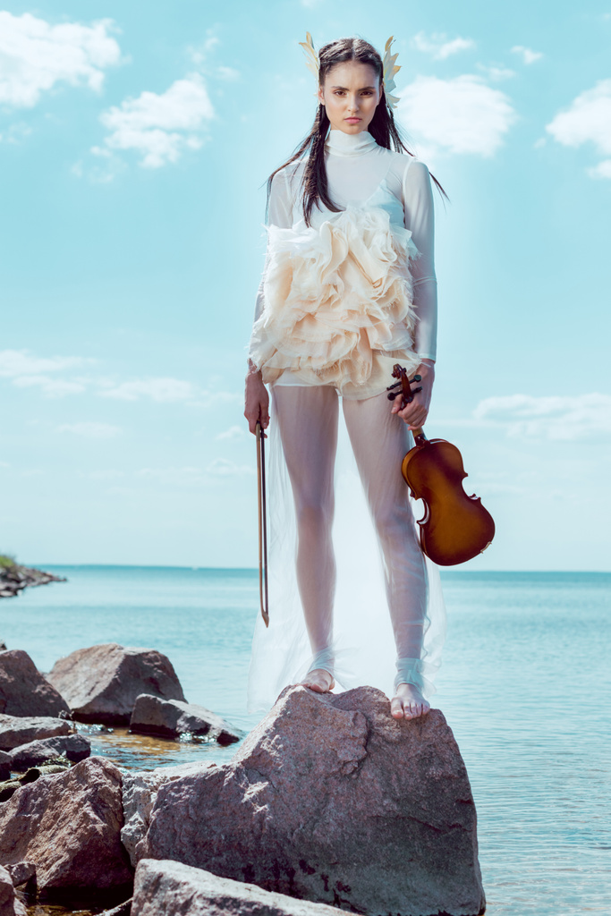 Tiefansicht einer eleganten Frau im weißen Schwanenkostüm mit Geige, die auf blauem Fluss und Himmelshintergrund steht - Foto, Bild