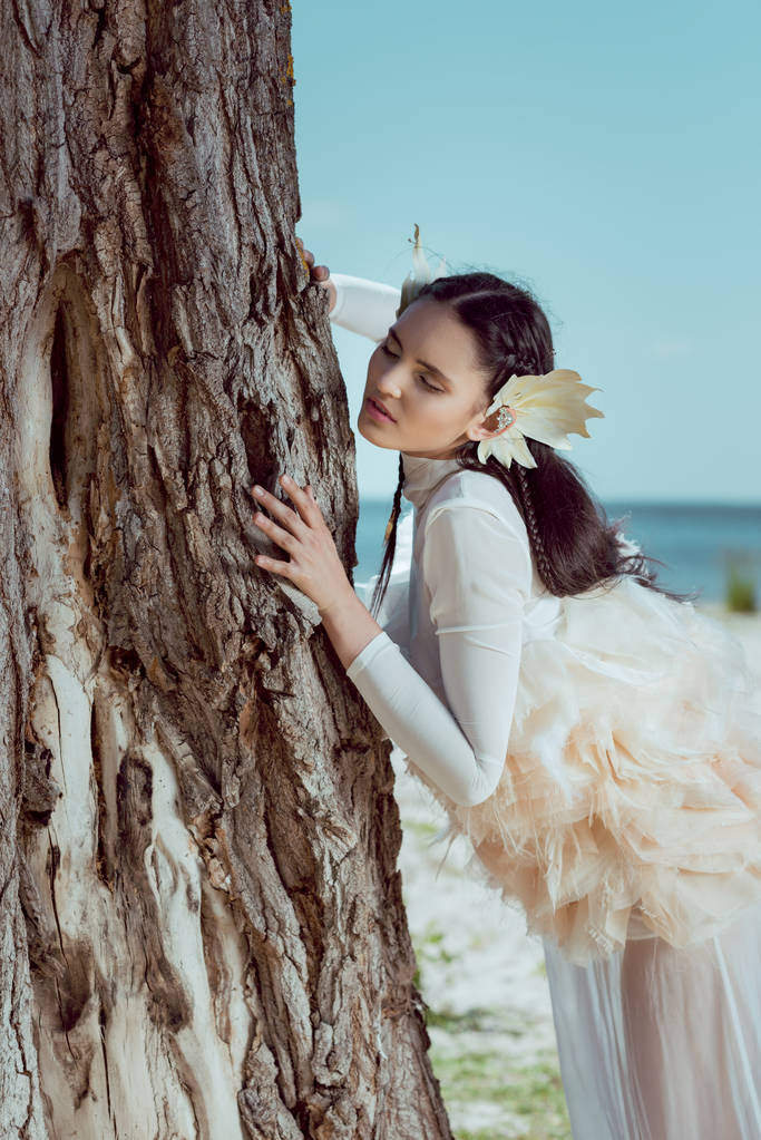 elegant woman in white swan costume standing near three trunk - Photo, Image