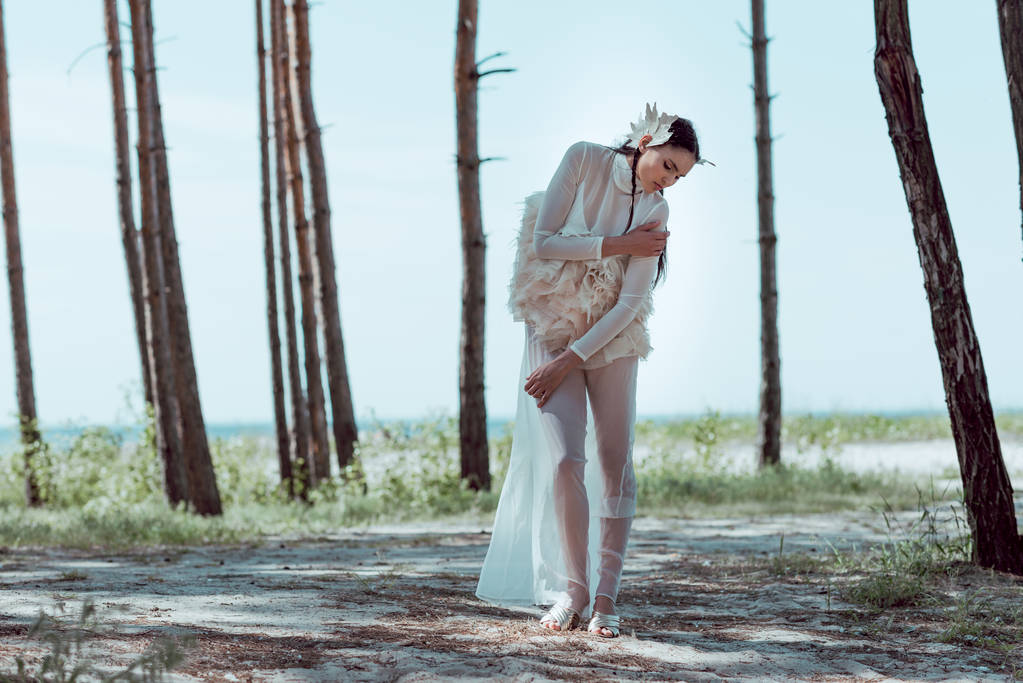beautiful woman in white swan costume standing on sandy beach near trees - Photo, Image