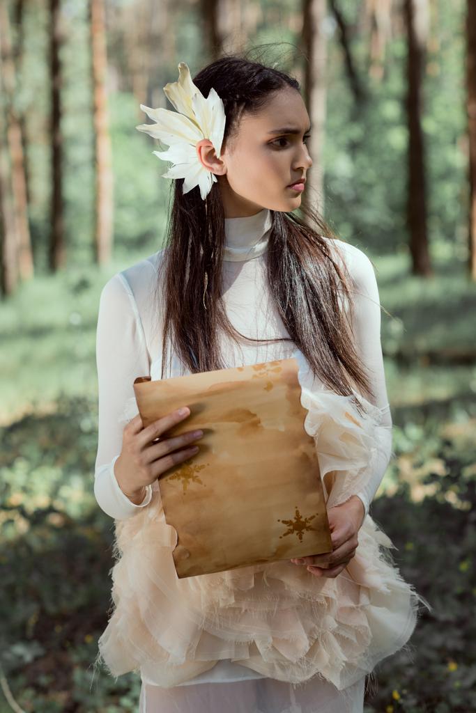 elegante donna in costume da cigno bianco con pergamena in piedi sullo sfondo della foresta, distogliendo lo sguardo
 - Foto, immagini