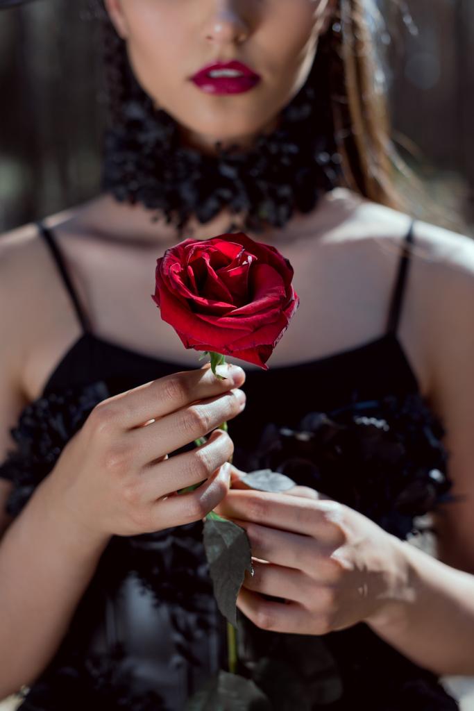 手に赤いバラを持つ魔女の衣装を着た若い女性のトリミングされたビュー ロイヤリティフリー写真 画像素材