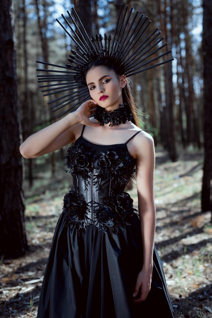 森の背景に立つ魔女の衣装でエレガントな女性の選択的な焦点, 首の近くに手を保持, カメラを見て - 写真・画像