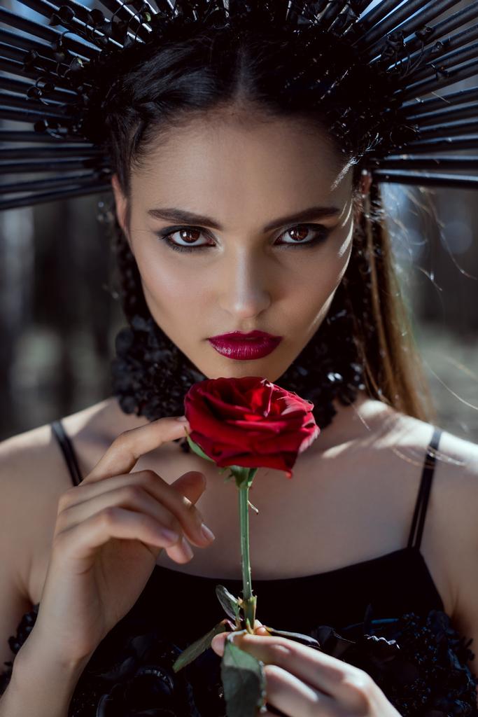 Porträt einer schönen Frau im Hexenkostüm, die in die Kamera blickt und eine rote Rose in den Händen hält - Foto, Bild