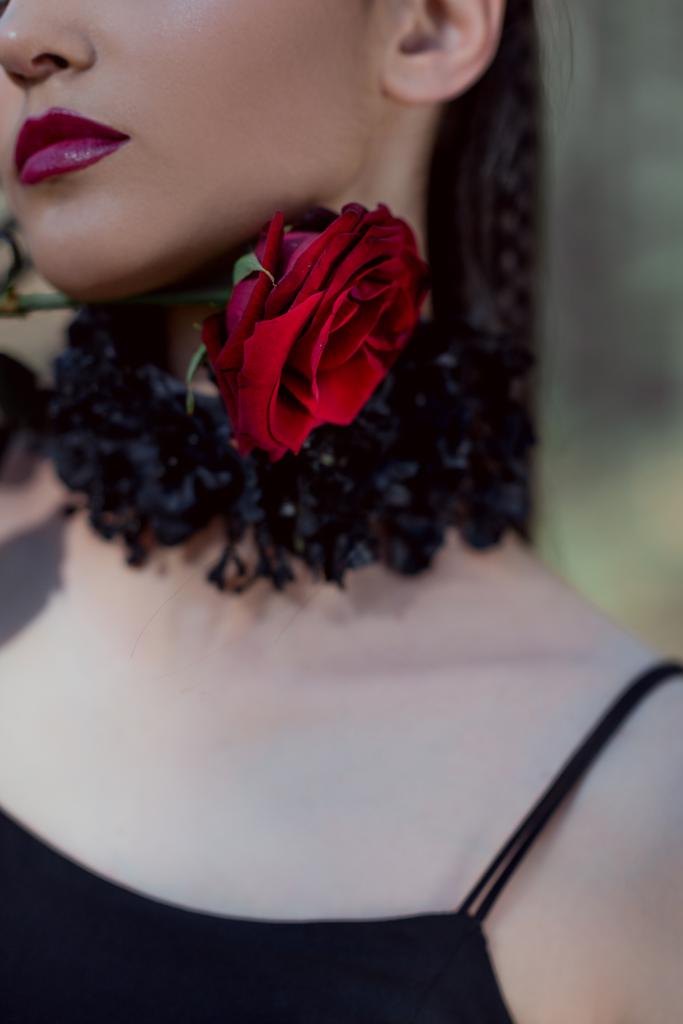 περικομμένη θέα της κομψής γυναίκας στο κοστούμι μάγισσα κρατώντας κόκκινο τριαντάφυλλο κοντά στο λαιμό - Φωτογραφία, εικόνα