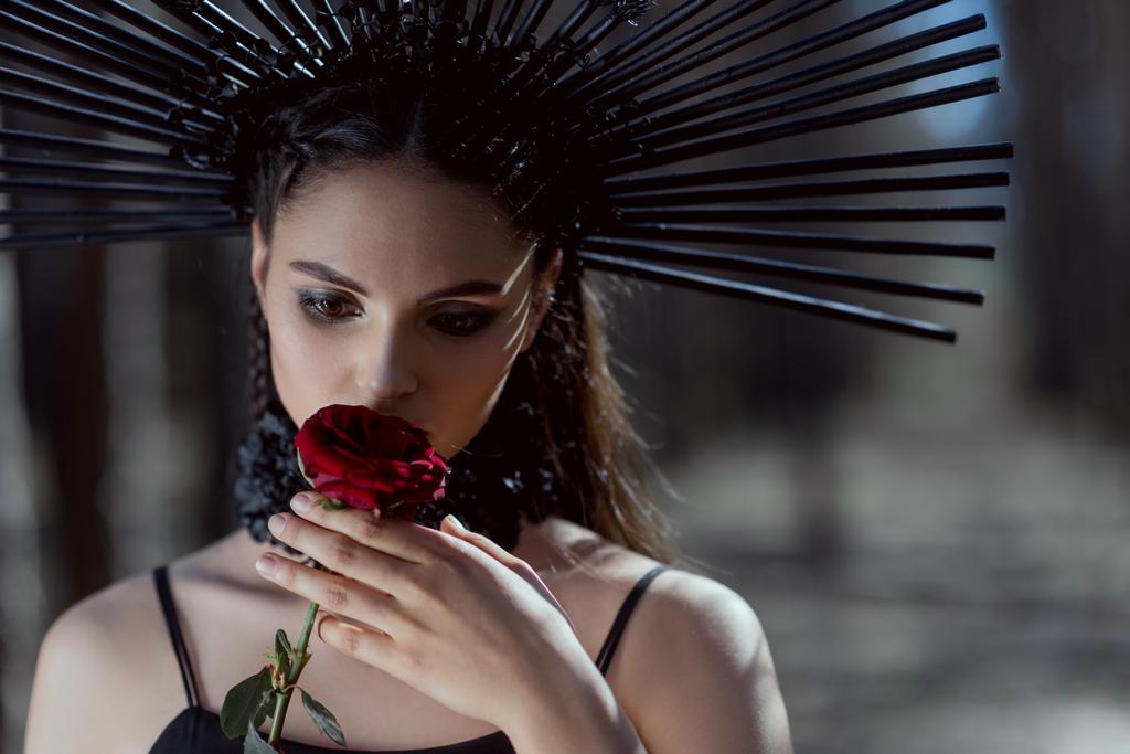 πορτραίτο της νεαρής γυναίκας με στολή μάγισσας με στέμμα στο κεφάλι κρατώντας κόκκινο τριαντάφυλλο κοντά στο πρόσωπο - Φωτογραφία, εικόνα