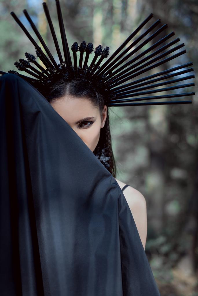 bela mulher em traje de bruxa com coroa na cabeça em pé atrás de têxteis pretos, olhando para a câmera
 - Foto, Imagem