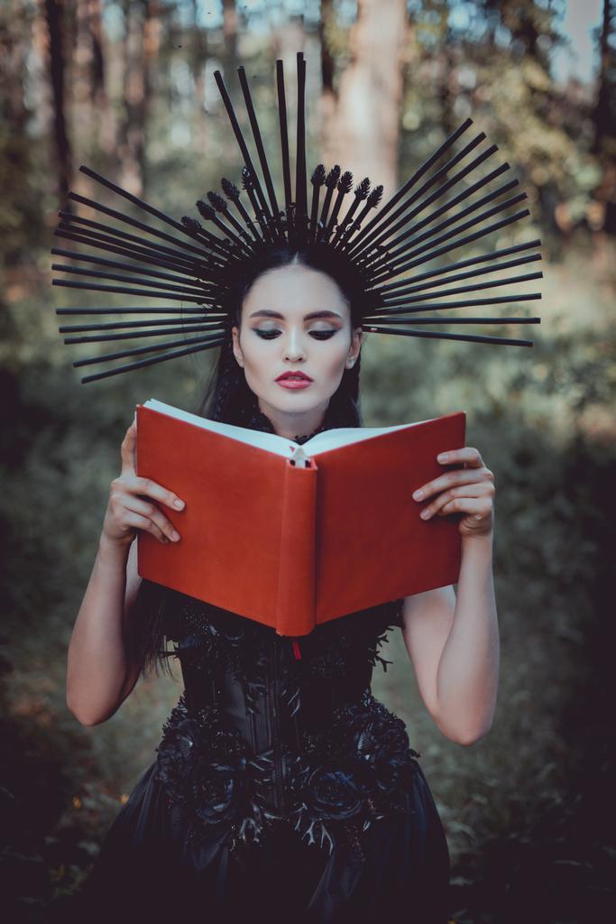 bela mulher no traje de bruxa em pé no fundo da floresta, lendo livro vermelho
 - Foto, Imagem