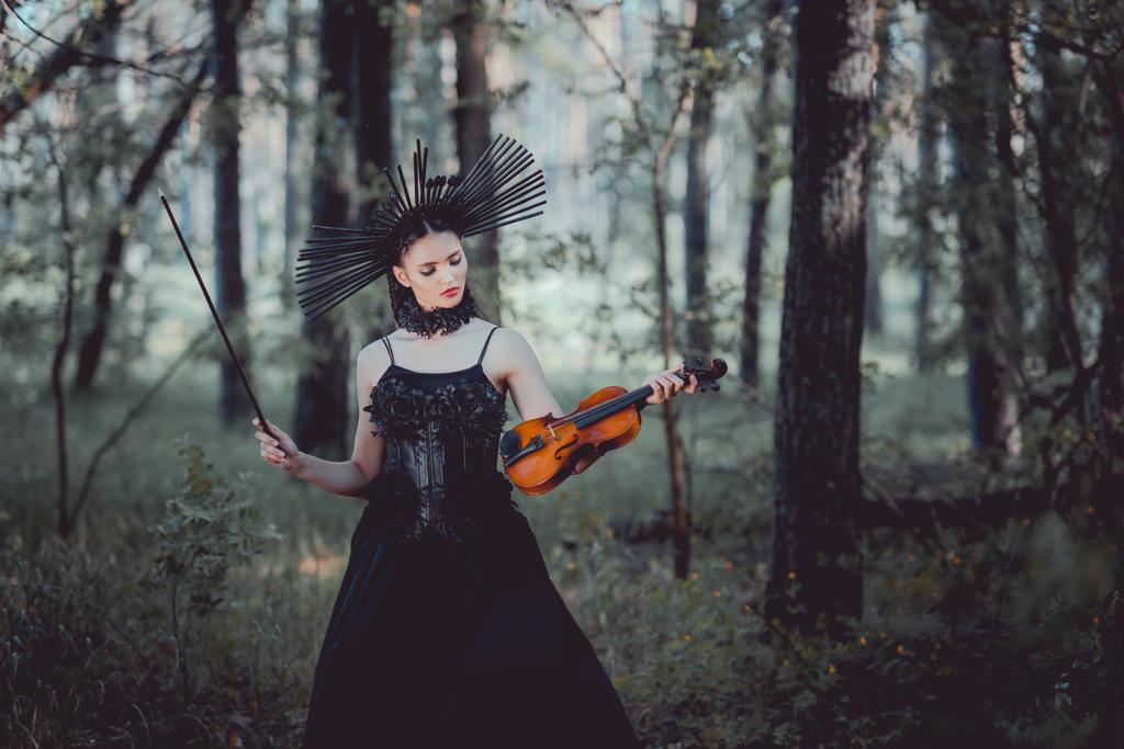 elegant woman in witch costume standing on forest background, looking at violin - Photo, Image