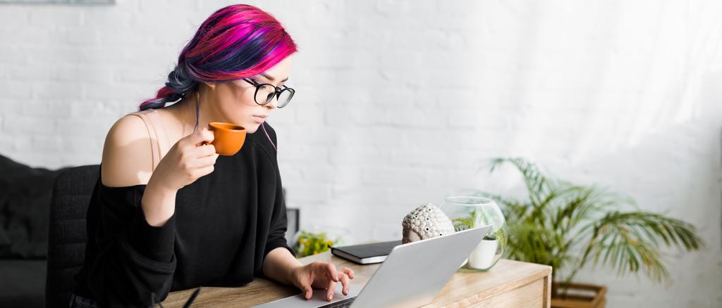 panoramic shot of beautiful girl with colorful hair drinking coffee while sitting at table and using laptop  - Photo, Image