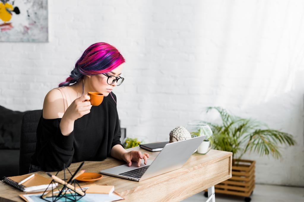  beautiful girl with colorful hair drinking coffee while sitting at table and using laptop  - Photo, Image