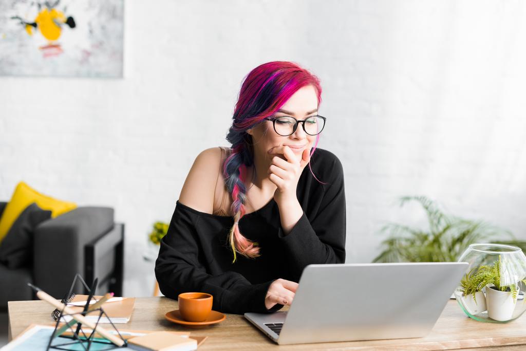 hipster girl with colorful hair sitting behind table and looking at laptop pensively - Photo, Image
