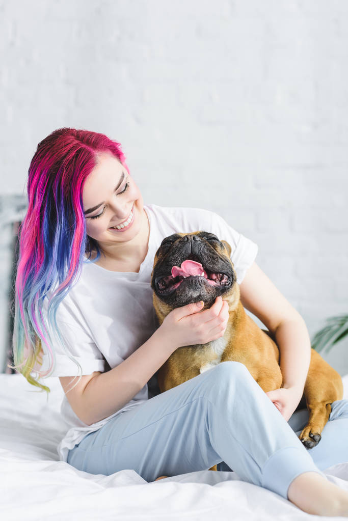 fröhliches Hipster-Mädchen sitzt auf dem Bett und streichelt französische Bulldogge  - Foto, Bild