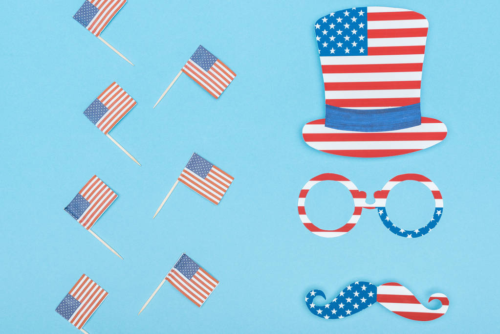 flat lay with hat, glasses and mustache made of stars and stripes near decorative american flags on wooden sticks on blue background - Photo, Image