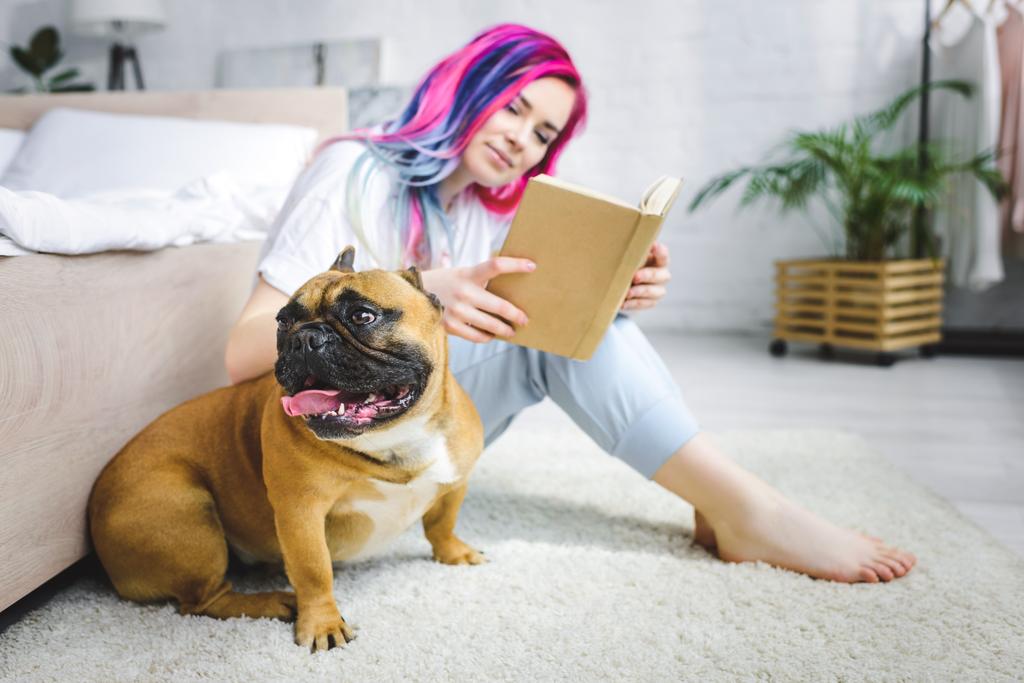 beautiful girl with colorful hair reading book and sitting on floor near French bulldog - Photo, Image