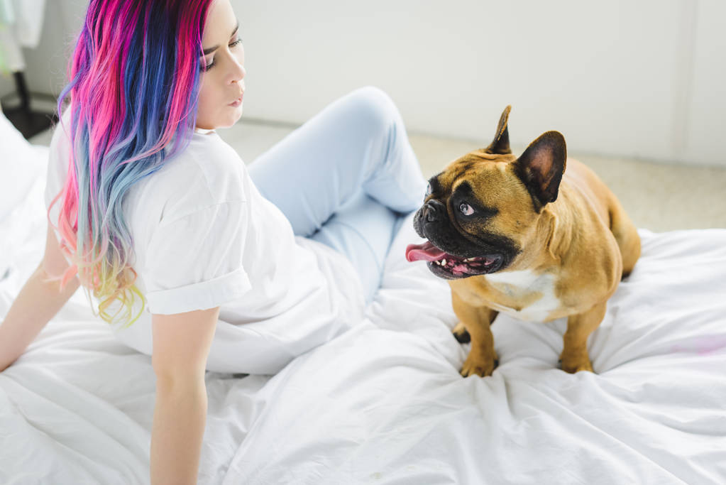 schönes Mädchen mit buntem Haar, das pfeift und die französische Bulldogge ansieht, während sie auf dem Bett sitzt  - Foto, Bild