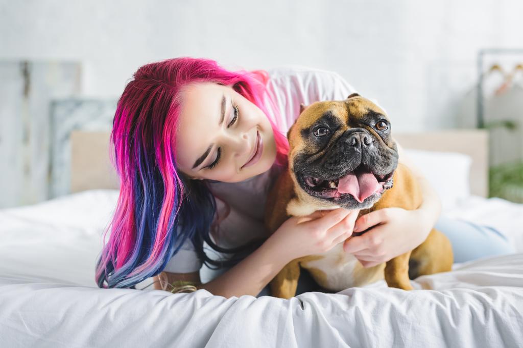 meisje met kleurrijke haren aaien en kijken naar schattige Franse Bulldog tijdens het leggen in bed - Foto, afbeelding