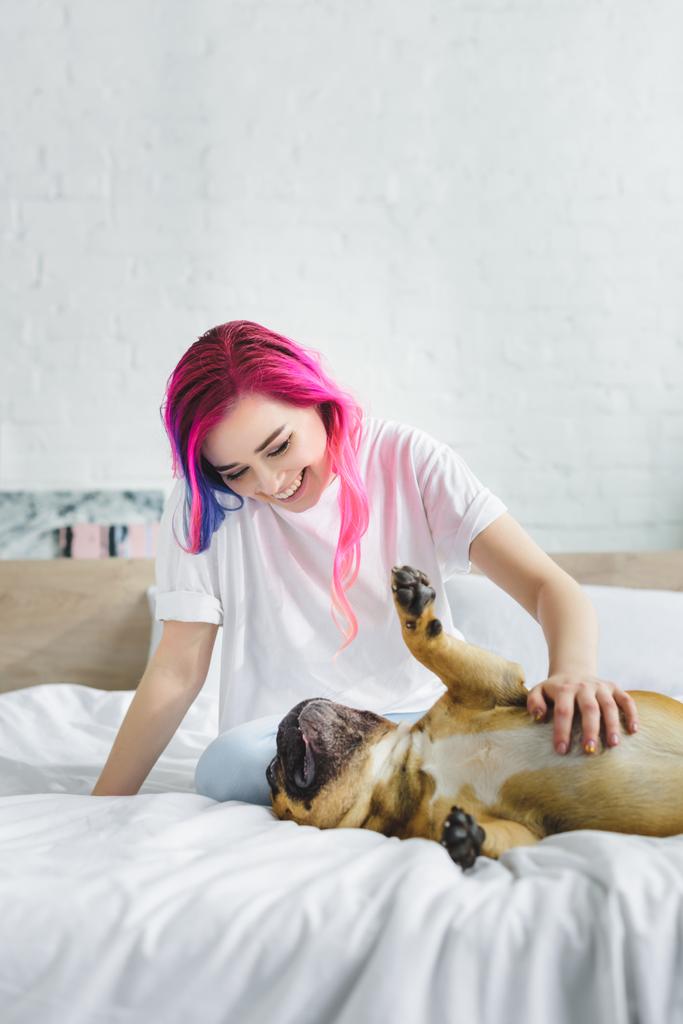 Mädchen mit bunten Haaren streichelt und schaut auf süße französische Bulldogge, die auf dem Rücken im Bett liegt - Foto, Bild