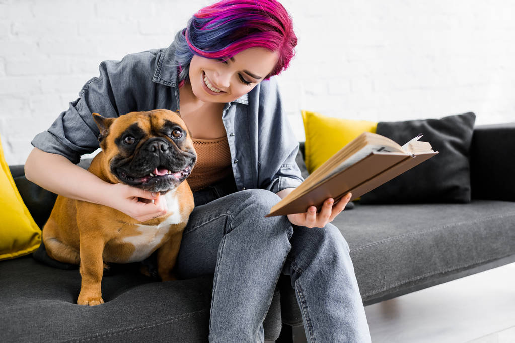 mooi meisje met kleurrijke haren knuffelen Bulldog, holding boek, glimlachend en zittend op de Bank - Foto, afbeelding