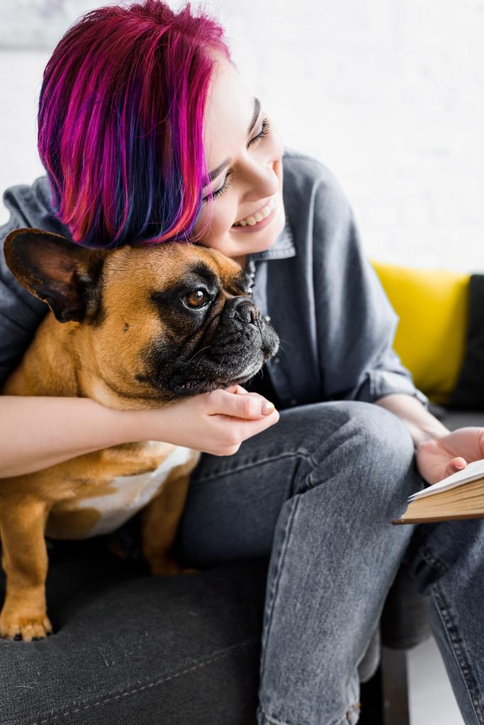 menina com cabelo colorido abraçando bulldog, sorrindo e segurando livro
  - Foto, Imagem