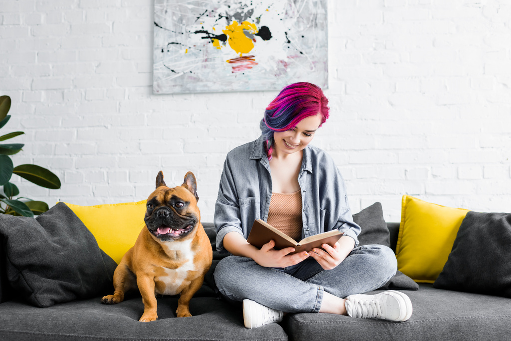 schönes Hipstermädchen sitzt auf Sofa neben kleiner französischer Bulldogge und liest Buch - Foto, Bild