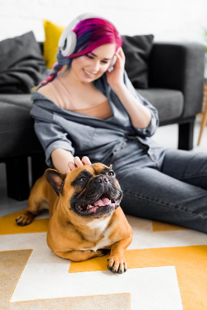 foco seletivo da menina bonita com cabelos coloridos e fones de ouvido sentado no chão e acariciando bulldog francês
 - Foto, Imagem