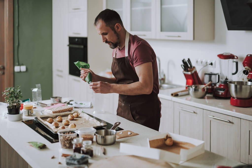 Hombre tranquilo de pie frente a la mesa de la cocina y poner crema en la pastelería
 - Foto, Imagen