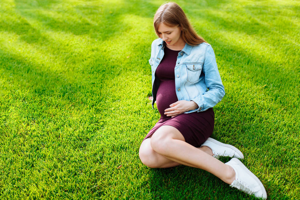 Šťastná Mladá těhotná žena, která sedí v parku na trávníku, - Fotografie, Obrázek