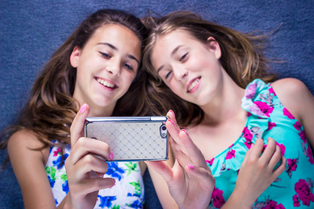 zwei Mädchen, die auf dem Bett liegen und auf ihr Smartphone schauen, Selfie machen und lächeln. - Foto, Bild