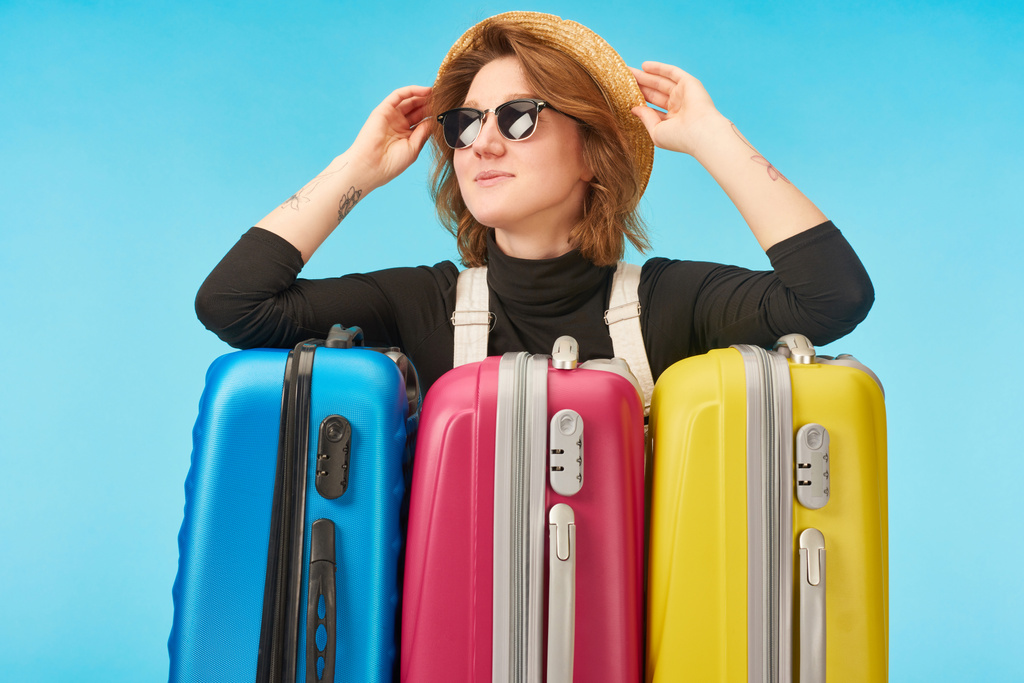 cheerful girl in sunglasses and straw hat near multicolored travel bags isolated on blue - Photo, Image