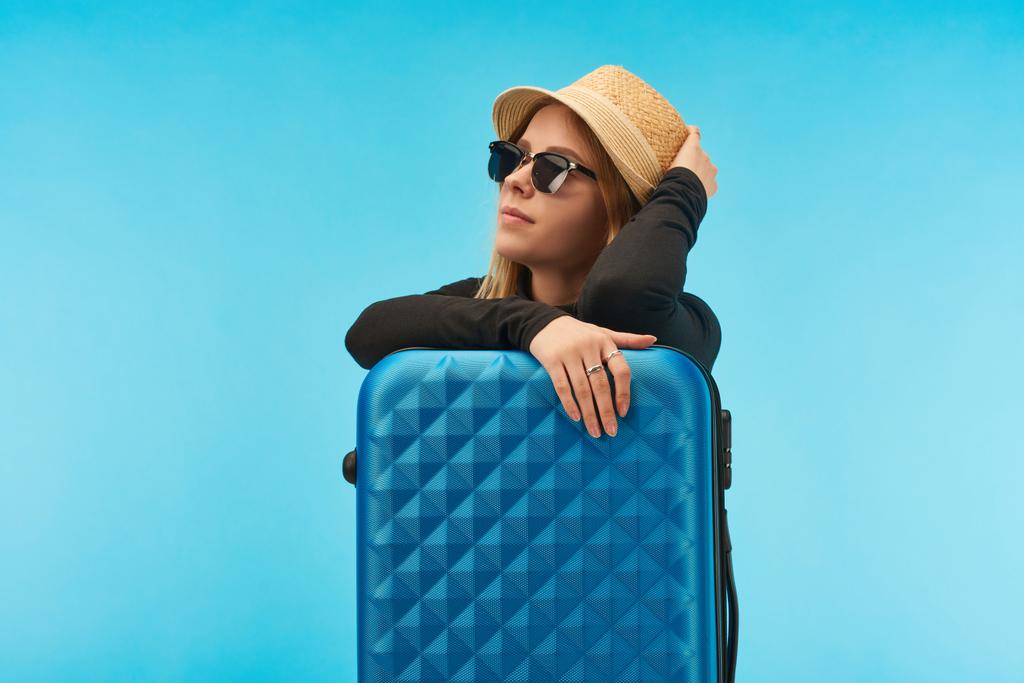 blonde girl in sunglasses and straw hat near blue suitcase isolated on blue - Photo, Image