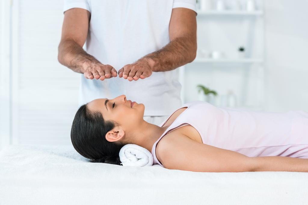 bijgesneden weergave van genezer zetten handen boven hoofd van aantrekkelijke vrouw met gesloten ogen liggend op massagetafel - Foto, afbeelding