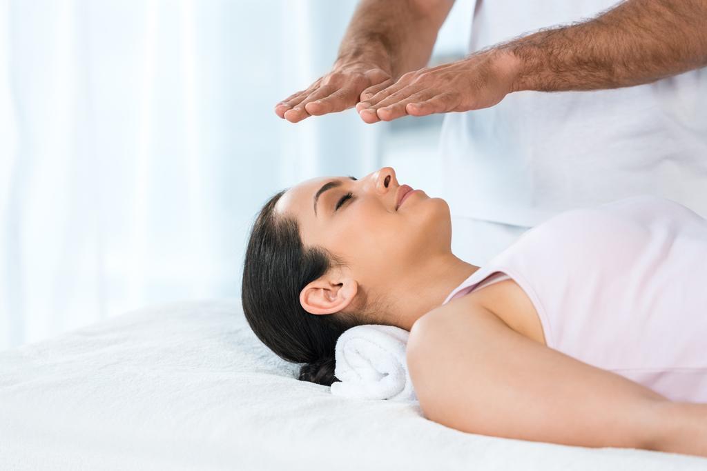 vue recadrée de l'homme mettant les mains au-dessus de la tête de la femme attrayante avec les yeux fermés couché sur la table de massage
 - Photo, image