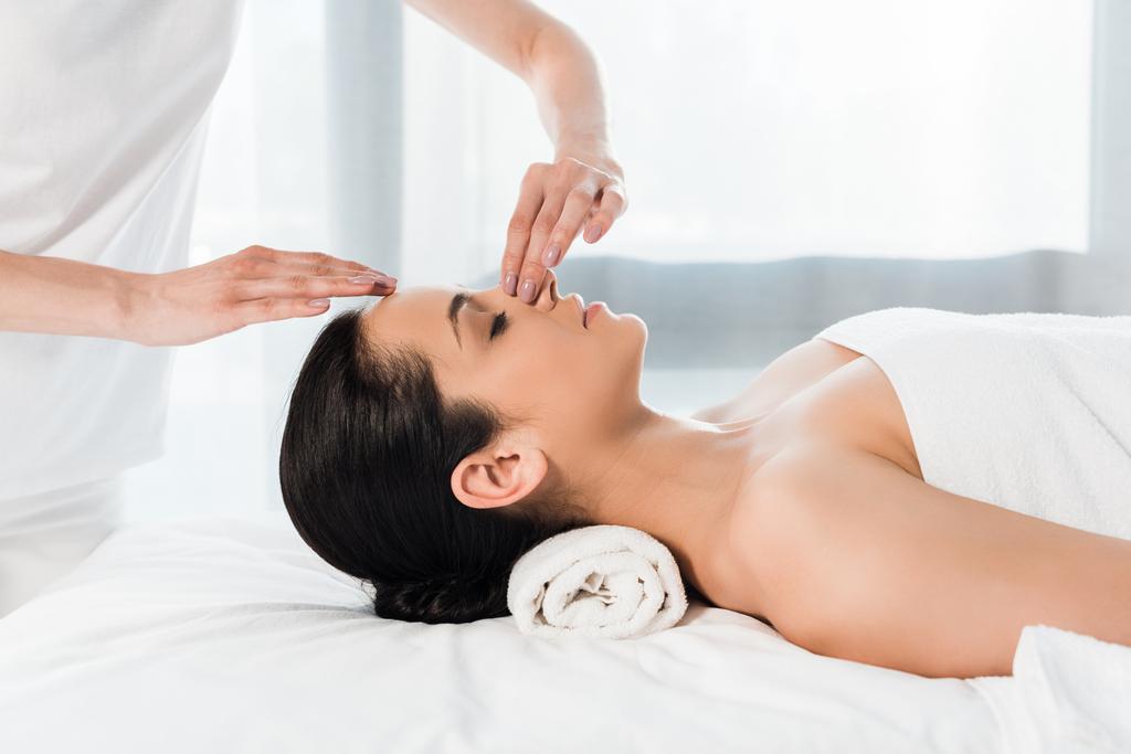 bijgesneden weergave van masseur aanraken neus van aantrekkelijke brunette vrouw liggend op massagetafel  - Foto, afbeelding