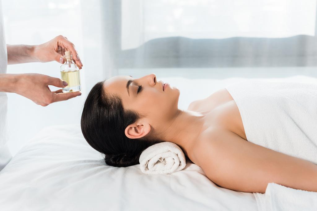 vue recadrée du masseur tenant bouteille avec de l'huile près de belle femme brune couchée sur la table de massage
  - Photo, image