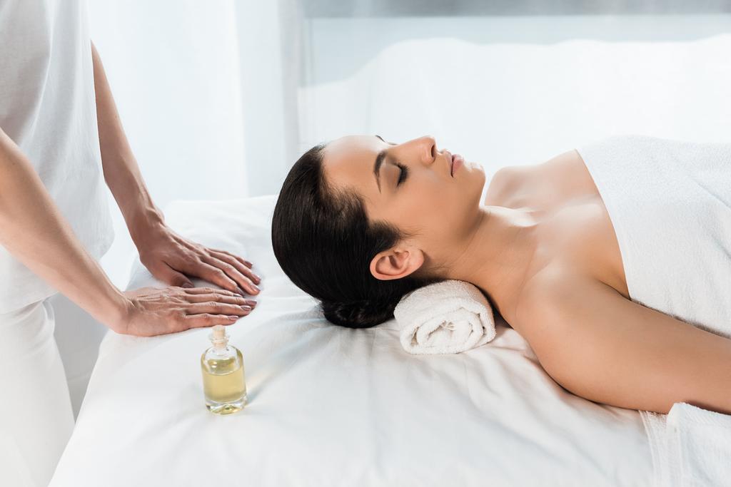 обрізаний вид на масажиста біля привабливої брюнетки з закритими очима, лежачи на масажному столі
  - Фото, зображення