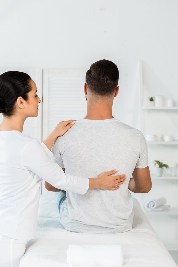 back view of man sitting on massage table near attractive healer  - Photo, Image