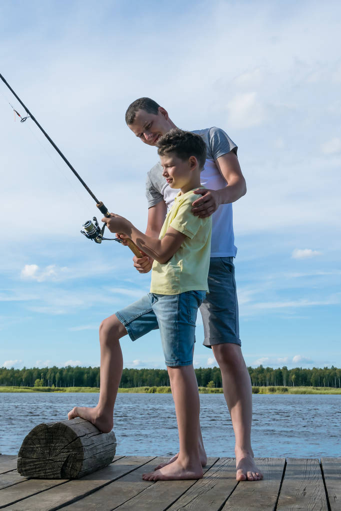 Isä opettaa poikaansa pyörittämään kunnolla kalastaessaan joella olevasta laiturista.
 - Valokuva, kuva