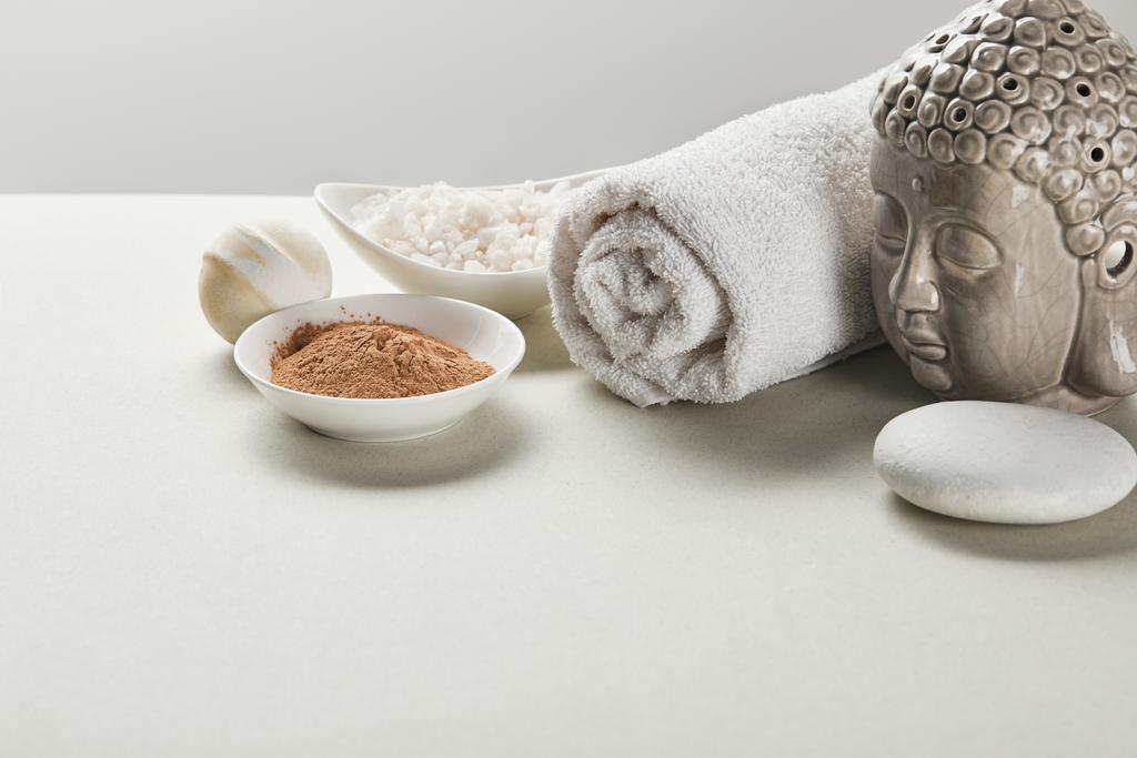 морская соль и глиняный порошок в мисках, хлопковое полотенце, камень, бомба для ванны и статуэтка Будды на белом столе, изолированном на сером
 - Фото, изображение
