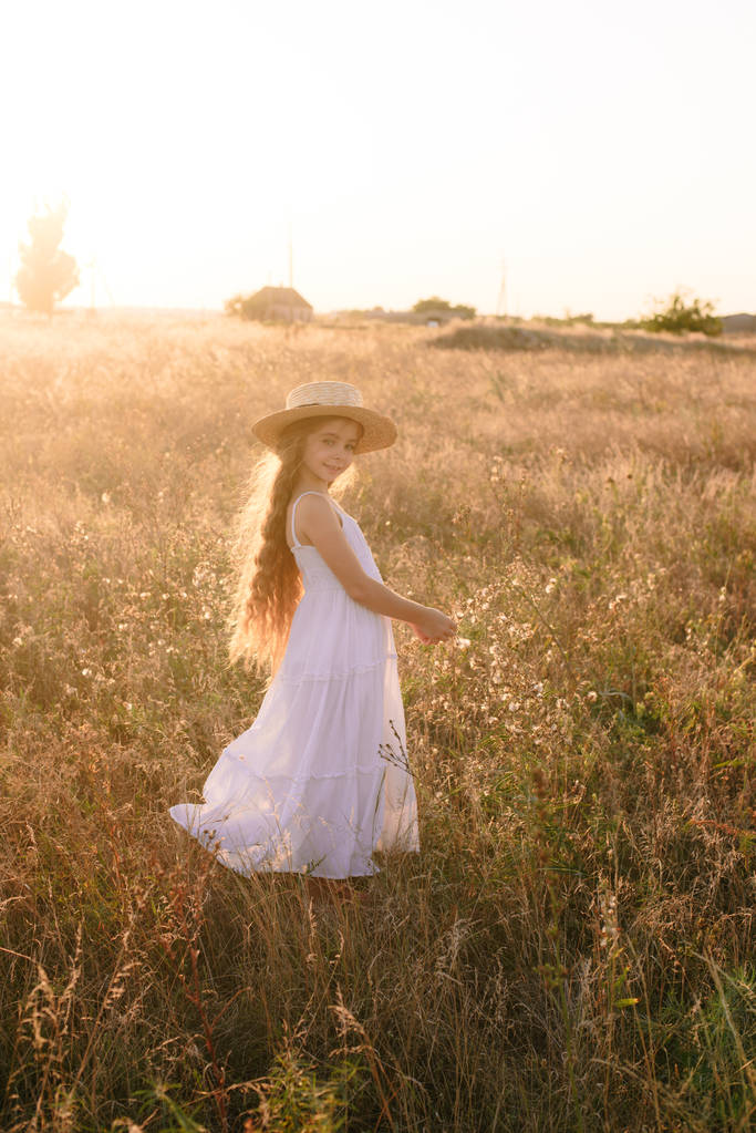 Söpö pieni tyttö vaaleat pitkät hiukset kesäkentällä auringonlaskun aikaan valkoinen mekko olki hattu
 - Valokuva, kuva