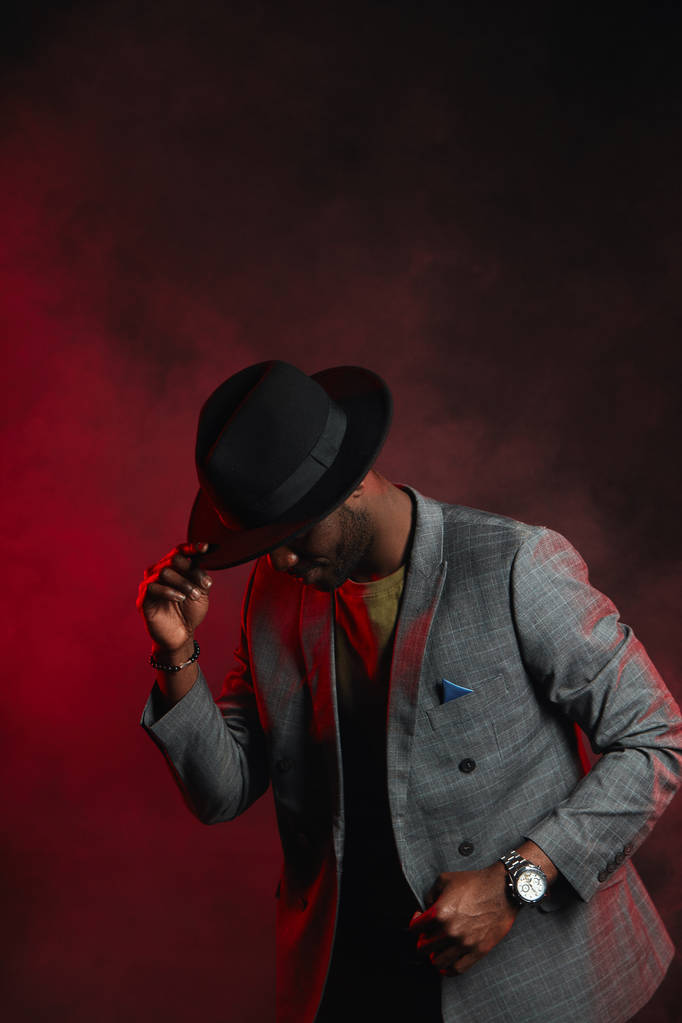 Afrikanischer Geschäftsmann in grauem Anzug und schwarzem Hut im Studio auf dunklem Hintergrund - Foto, Bild