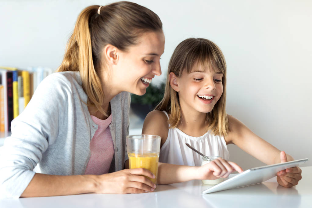 Όμορφη νεαρή μητέρα και η κόρη της χρησιμοποιώντας το ψηφιακό δισκίο, ενώ το πρωινό στο σπίτι.  - Φωτογραφία, εικόνα