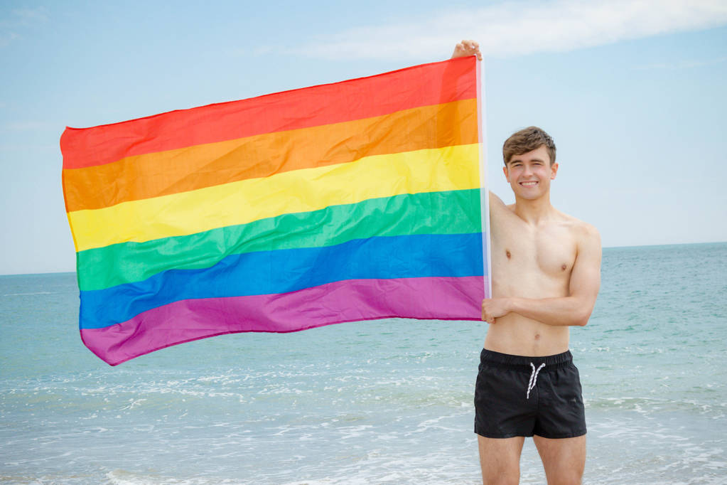 Белый мужчина на пляже с флагом Прайда
 - Фото, изображение