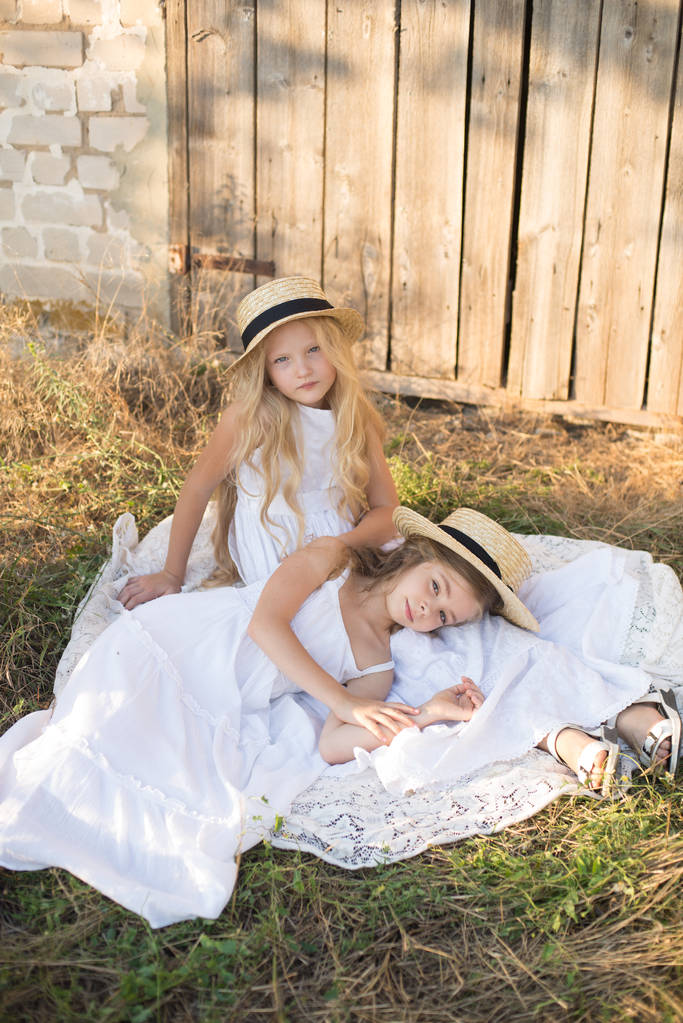 Lindas niñas con el pelo largo y rubio en un campo de verano al atardecer en vestidos blancos con un sombrero de paja
 - Foto, imagen