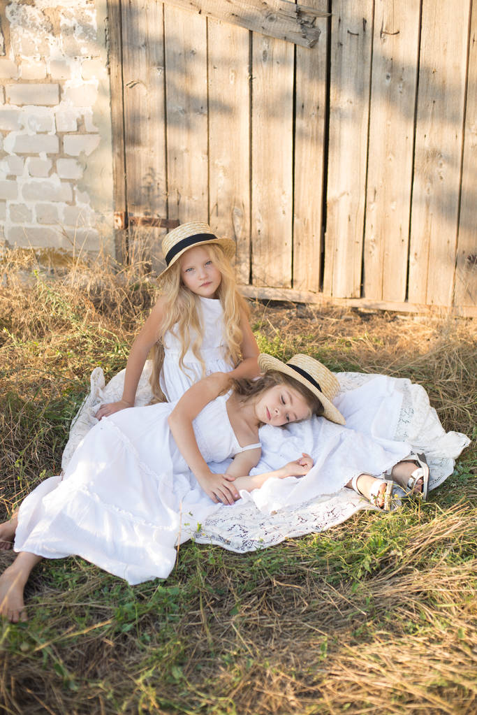 Χαριτωμένα μικρά κορίτσια με ξανθά μακριά μαλλιά σε ένα καλοκαιρινό χωράφι στο ηλιοβασίλεμα με λευκά φορέματα με ψάθινο καπέλο - Φωτογραφία, εικόνα
