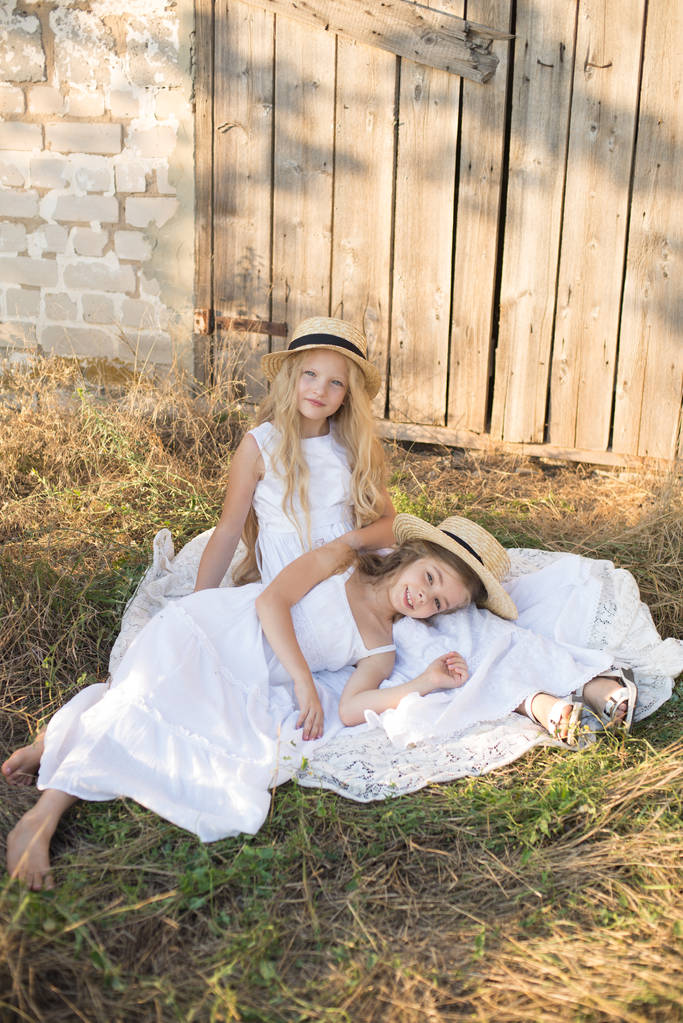 Lindas niñas con el pelo largo y rubio en un campo de verano al atardecer en vestidos blancos con un sombrero de paja
 - Foto, imagen