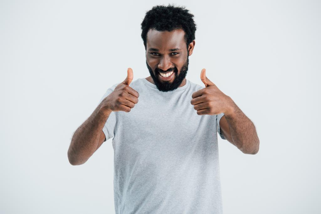 улыбающийся африканский американец в серой футболке, показывающий большие пальцы, изолированные на сером
 - Фото, изображение