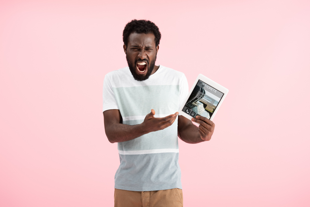 感情的なアフリカ系アメリカ人男性が悲鳴を上げ、チケットアプリでデジタルタブレットを示し、ピンクで隔離 - 写真・画像