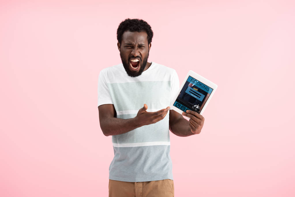 συναισθηματικός Αφρικανός Αμερικανός άνθρωπος ουρλιάζοντας και δείχνοντας ψηφιακό δισκίο με την εφαρμογή κρατήσεων, απομονώνονται σε ροζ - Φωτογραφία, εικόνα